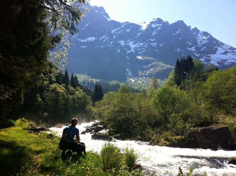 A hiker resting by a rushing mountain river below snow-capped peaks.